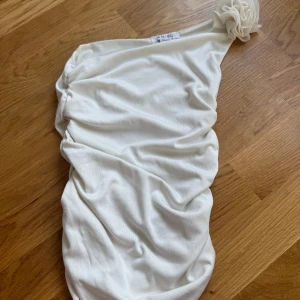 Vit oneshoulder topp - Säljer en stilren vit oneshoulder topp med en dekorativ blomdetalj på axeln. Klänningen har en tight passform som framhäver figuren. Perfekt för en elegant kväll.