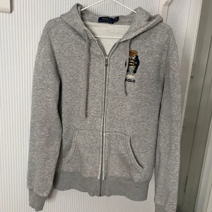 Grå zip-hoodie från Polo Ralph Lauren - Säljer en stilren grå hoodie från Polo Ralph Lauren med dragkedja och en broderad björn på bröstet. Tröjan har en klassisk design med fickor framtill och ribbade muddar vid ärmslut och nederkant. På västerärm har den en liten defekt, se näst sista bilden. Tveka inte att höra av dig vid intresse eller frågor!