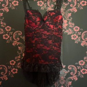 Svart och röd korsett från Sexkiss - Snygg svart och röd korsett från Sexkiss med spetsdetaljer och justerbara axelband. Den har en elegant design med volangdetaljer och en öppen rygg. 