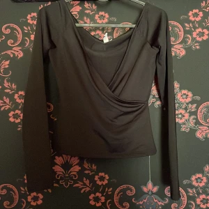 Svart omlottopp från Nelly - Snygg svart omlottopp från Nelly med långa ärmar och en elegant v-ringning. Perfekt för en stilren look. Toppen är i en mjuk och bekväm polyesterblandning. använd en gång