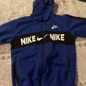 Blå hoodie från Nike - Säljer en blå hoodie från Nike med dragkedja och huva. Jackan har ett stort Nike-tryck i vitt på bröstet och svarta detaljer längs ärmarna. Perfekt för en sportig look!