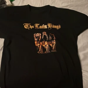 Svart T-shirt med The Latin Kings-tryck - Cool svart T-shirt med tryck av The Latin Kings i guld och en bild på framsidan. Perfekt för fans av bandet eller för en avslappnad stil.