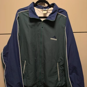 Classic vindjacka från Adidas - Säljer en grön och blå vindjacka från Adidas med dragkedja och vita detaljer. Jackan har långa ärmar och två fickor framtill. Perfekt för en sportig look. 