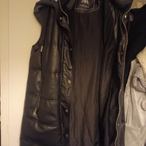 Svart dunväst från Zara - Snygg svart dunväst från Zara med dragkedja och knappar. Perfekt för kyligare dagar med sin quiltade design och huva. Västen har praktiska fickor och en stilren look.