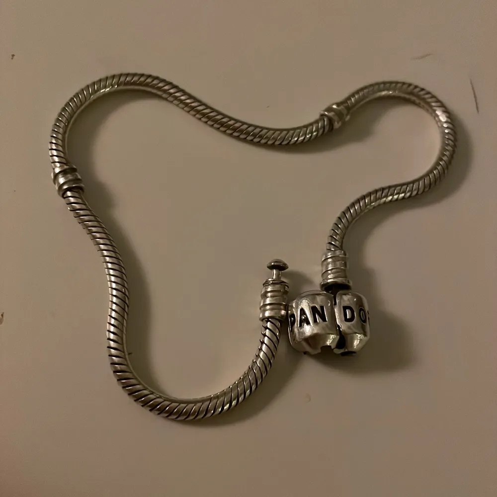 Säljer ett elegant silverfärgat armband från Pandora. Armbandet har en klassisk ormkedjedesign med en rund låsdetalj där Pandora-loggan är ingraverad. Perfekt för att fästa dina favoritberlocker och skapa en personlig stil.. Asusteet.