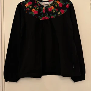Svart tröja med blommigt mönster från Polarn O. Pyret - Säljer en svart långärmad tröja från Polarn O. Pyret med ett färgglatt blommigt mönster på axlarna och runt halsen. Perfekt för att liva upp garderoben med lite färg och mönster! Storlek 146/152. Följer med en gratis liten knapp! 