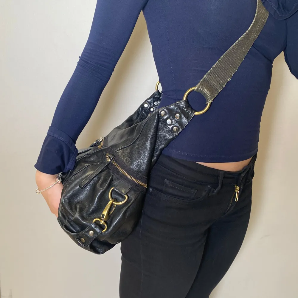 Snygg svart axelväska i skinn med coola nitar och dragkedjedetaljer. Perfekt för att ge din outfit en edgy touch. Väskan har en justerbar axelrem och flera praktiska fack för enkel organisering.. Laukut & Käsilaukut.
