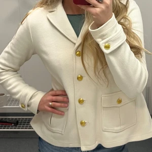 Vit kavaj med guldknappar - Snygg vit kavaj/tröja i stickat material med guldknappar och två framfickor. Kavajen har en klassisk krage och långa ärmar, perfekt för en stilren look.