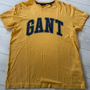 Gul t-shirt från GANT strl M - Snygg gul t-shirt från GANT med stort tryck i marinblått på framsidan. Perfekt för en avslappnad stil. Från djur och rökfritt hem