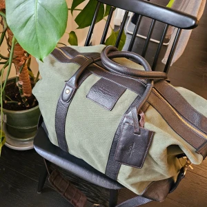 Grön och brun väska med detaljer - Snygg grön väska med bruna läderdetaljer och dragkedja. Perfekt för resor eller som en rymlig vardagsväska. Den har robusta handtag och metallfötter för extra hållbarhet.