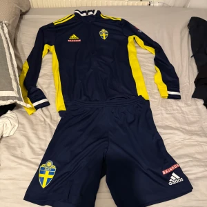 Landslags kofta och shorts  - Säljer en blå och gul långärmad fotbollströja med matchande shorts från Adidas. Tröjan har Sveriges landslagsemblem på bröstet och gula detaljer längs ärmarna. Perfekt för fotbollsfans som vill visa sitt stöd. Pris kan diskuteras