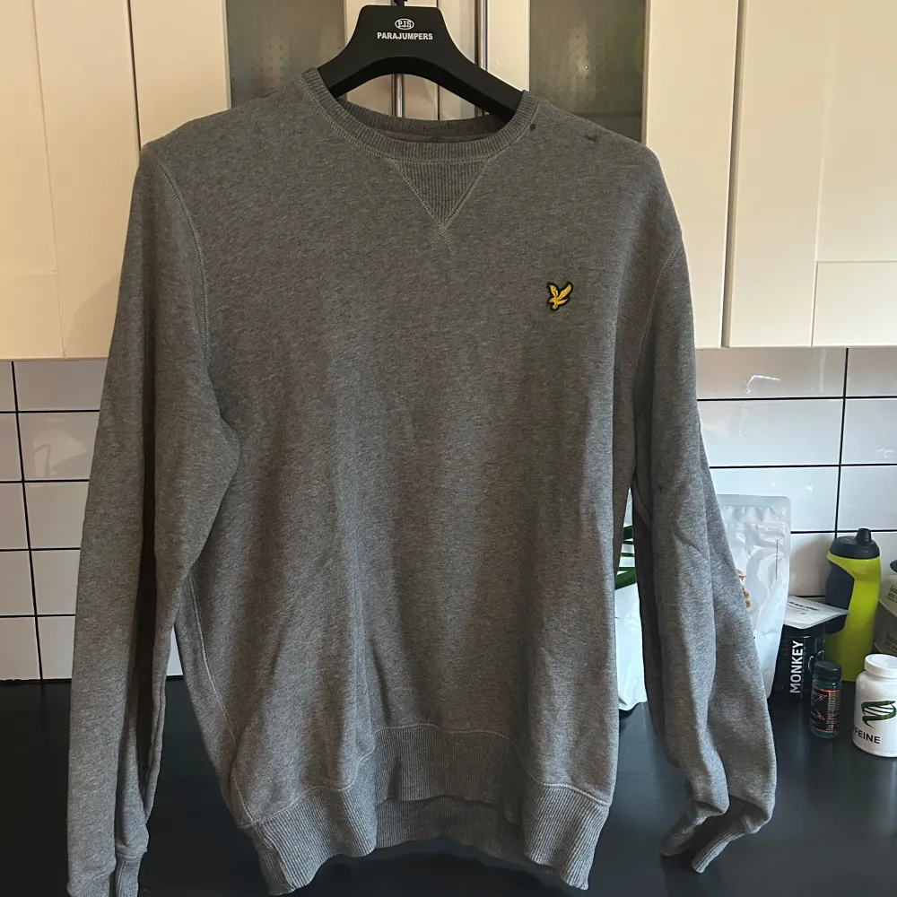Säljer en stilren grå tröja från Lyle & Scott med rund hals och ribbade muddar. Tröjan har en liten broderad logotyp på bröstet. Perfekt för en avslappnad look.. Neuletakit & Villapaidat.