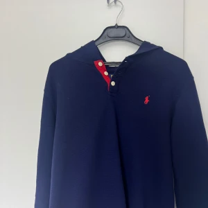 Mörkblå hoodie från Ralph Lauren - Säljer en snygg mörkblå hoodie från Ralph Lauren med röda detaljer och knappar. Den har en klassisk design med lång ärm och en liten logga på bröstet. Perfekt för en stilren look.