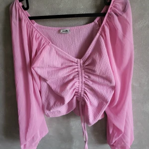 Rosa topp från Pimkie - Säljer en söt rosa topp från Pimkie. Toppen har långa ärmar och en snygg knytdetalj framtill som ger en rynkad effekt. Perfekt för en somrig look!