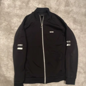 Svart jacka från Hugo Boss - Snygg svart jacka från Hugo Boss med dragkedja framtill och ribbad krage. Jackan har märkets logga på bröstet och ärmarna, vilket ger en stilren look. Perfekt för en casual stil.