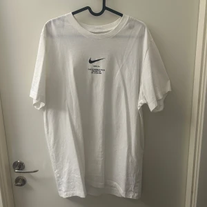 Vit t-shirt från Nike - Använd 1 gång. Snygg vit t-shirt från Nike med svart logga och text på både fram- och baksidan. Perfekt för en sportig look eller avslappnad stil. Tillverkad i mjukt material för extra komfort.