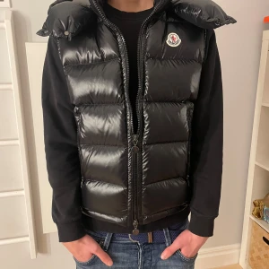 Svart dunväst från Moncler - Säljer en snygg svart dunväst från Moncler med dragkedja och huva. Västen har en quiltad design och en broderad logotyp på bröstet. Perfekt för kyligare dagar. Priset kan sänkas vid snabb o seriös affär 👍