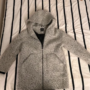 Grå kappa från Clockhouse /Storlek XL - Säljer en stilren grå kappa från Clockhouse med huva och dragkedja. Jackan har en mjuk och mysig textur, perfekt för kyligare dagar. Den har praktiska fickor framtill och en klassisk passform.