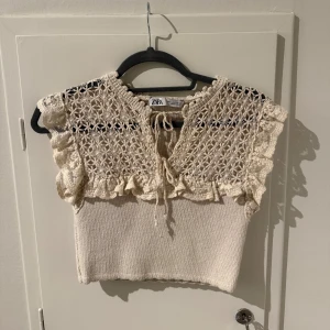 Beige stickad topp från Zara - Säljer en söt beige stickad topp från Zara med vackra virkade detaljer och volanger. Toppen har en knytning framtill och är ärmlös, perfekt för varmare dagar. Den har en croppad passform som ger en trendig look, bara att höra av dig vid frågor💕