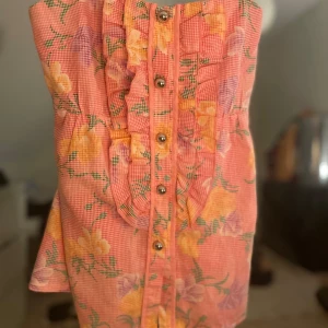 Blommig topp med volangdetaljer - Charmig blommig topp i rosa med gula och lila blommor. Klänningen har smala axelband och knappar framtill. Volangdetaljer pryder framsidan och ger en romantisk touch. den har använts 4/6 gånger