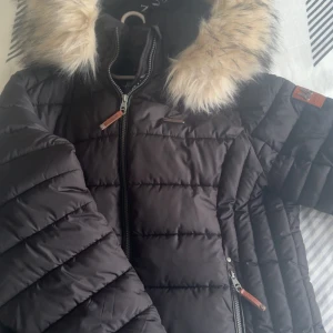 Svart dunjacka från Marikoo - Snygg svart dunjacka från Marikoo med pälskantad huva. Jackan har dragkedja och quiltad design, perfekt för kalla dagar. Den har en stilren look med detaljer i brunt läder vid dragkedjorna.
