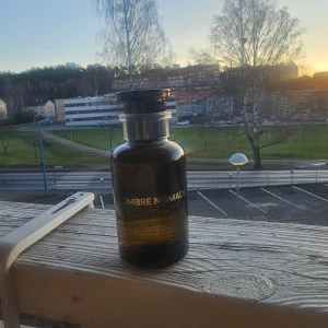 Ombre Nomade parfym från Louis Vuitton - Elegant parfymflaska i mörkt glas med texten 'Ombre Nomade' i guld. Flaskan har en stilren design med en svart kork. Perfekt för den som söker en lyxig doftupplevelse.helt nytt