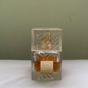Khamrah Parfym - Elegant parfym från Lattafa i en lyxig glasflaska med detaljerat mönster. Doften är varm och kryddig, perfekt för den som vill sticka ut. Flaskan har en fyrkantig form med en genomskinlig kork och en gyllene etikett framtill.