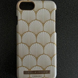 Mobilskal från Ideal of Sweden - Snyggt mobilskal från Ideal of Sweden med ett elegant mönster i vitt och guld. Skalet har en stilren design med ett skalformat mönster som ger en lyxig känsla. Perfekt för att skydda din telefon med stil.