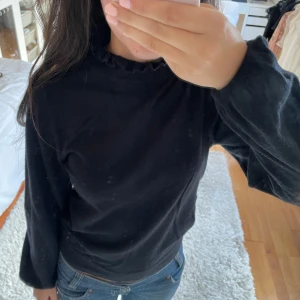 Svart långärmad topp med volangdetalj - Säljer en stilren svart långärmad topp med volangdetalj vid halsen. Perfekt för en fin och enkel look. Passar bra till jeans eller kjol.
