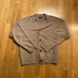 Beige kofta från Jack & Jones - Säljer en stilren beige kofta från Jack & Jones. Koftan har en v-ringad design med knappar framtill och ribbade muddar vid ärmslut och nederkant. Perfekt för lager-på-lager under kyliga dagar.
