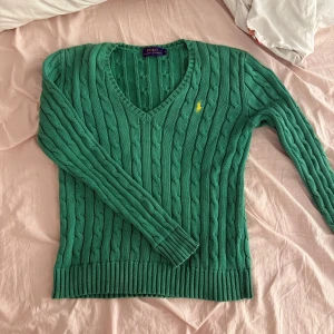 Grön stickad tröja från Ralph Lauren - Säljer en snygg grön stickad tröja från Ralph Lauren med kabelstickat mönster och v-ringning. Tröjan har långa ärmar och en liten gul logga på bröstet. Perfekt för en stilren look! Sparsamt använd men använd ganska mycket 🫶🏻jag bär vanligtvis storlek 38🫶🏻