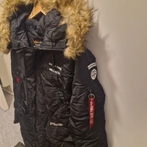 Svart parkajacka från Alpha Industries - Säljer en svart parkajacka från Alpha Industries med fuskpäls på huvan. Jackan har flera detaljer som knappar och dragkedja, samt broderade märken på bröstet och ärmen. Perfekt för kalla dagar med sin quiltade insida och praktiska fickor.