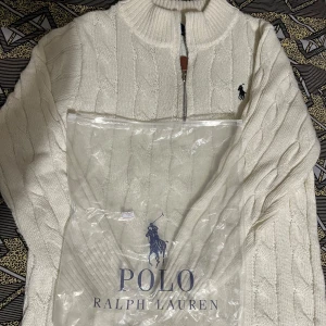 Vit stickad tröja från Ralph Lauren - Säljer en stilren vit stickad tröja från Ralph Lauren med dragkedja vid halsen. Tröjan har ett klassiskt flätmönster och långa ärmar. Perfekt för kyligare dagar. Storlek S&M