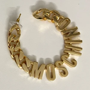 Guldörhängen från Moschino - Stilrena guldörhängen från Moschino med kedjedesign och bokstäver som bildar varumärkets namn. Perfekta för att ge en lyxig touch till din outfit. Aldrig använda. 