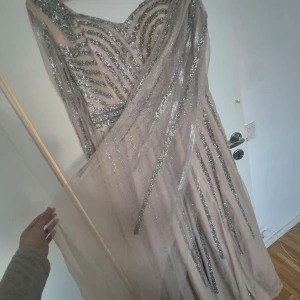 Glittrig långklänning med silverdetaljer - Elegant långklänning i ljus beige med glittriga silverdetaljer. Klänningen har en v-ringad design och långa ärmar med ett transparent utseende. Perfekt för festliga tillfällen med sin glamorösa stil.
