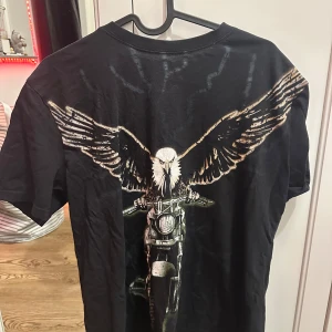 Svart t-shirt med örntryck - Cool svart t-shirt med ett stort tryck av en örn på en motorcykel . Perfekt för en avslappnad stil med en edgy touch.