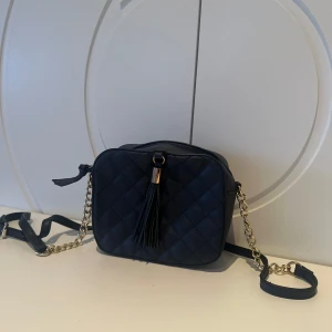 Marinblå quiltad axelväska  - Snygg marinblå axelväska med quiltat mönster och en dekorativ frans på framsidan. Väskan har en kedja som axelrem och stängs med dragkedja. Perfekt för att bära dina nödvändigheter med stil. Är marinblå men ser svart ut på bilden