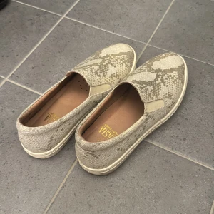 Beige slip-on skor från Dasia - Säljer ett par snygga och bekväma slip-on skor från Dasia i storlek 37. Skorna har ett coolt ormmönster i beige och grå toner, perfekt för att ge din outfit en extra touch. De är i bra skick och har en skön passform. Perfekta för vår och sommar!