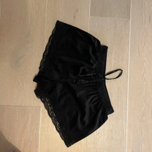 Svarta shorts med spets från Hunkemöller - Säljer ett par snygga och bekväma svarta shorts från Hunkemöller. De är gjorda i ett mjukt material och har fina spetsdetaljer längs kanterna. Perfekta för en avslappnad dag hemma eller som en del av en mysig outfit. Midjan är elastisk för extra komfort.