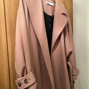 Beige kappa från Zara - Säljer en superfin beige kappa från Zara. Den är i mycket bra skick och perfekt för höst och vår. Kappan har långa ärmar och snygga knappar vid ärmsluten. Den är gjord i ett mjukt material som känns lyxigt. Passar perfekt för en stilren look! En söm har lossnat som håller bältet men går att fixa lätt. 