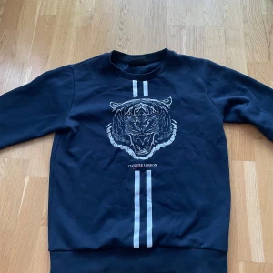 Svart sweatshirt från Closure London - Säljer en svart sweatshirt från Closure London med ett coolt tigertryck på framsidan. Tröjan har långa ärmar och två vita ränder som går vertikalt över bröstet. Perfekt för en avslappnad stil eller en dag ute med vänner. 🐯