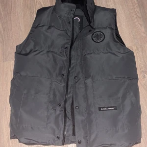 Grå dunväst från Canada Goose - Säljer en snygg grå dunväst från Canada Goose i storlek L. Västen är i mycket bra skick och har en dragkedja och knappar framtill samt två praktiska fickor. Perfekt för kyliga höst- och vinterdagar. Märket är broderat på bröstet och etiketten syns tydligt på framsidan. Passar både till vardags och lite finare tillfällen. Finns att hämta i Norrköping, pris kan diskuteras!