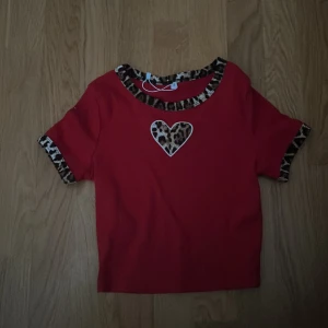 Röd topp med leoparddetaljer - Säljer en superfin röd topp med korta ärmar och leopardmönstrade detaljer vid hals och ärmslut. Den har ett sött leopardmönstrat hjärta på bröstet. Perfekt för en cool och avslappnad stil!