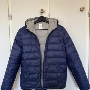 Mörkblå dunjacka med huva - Säljer en snygg mörkblå dunjacka från Polo Ralph Lauren. Jackan har en praktisk huva och dragkedja framtill. Perfekt för kyliga höst- och vinterdagar. Den är i mycket bra skick och har en stilren design med märkets logga på bröstet.