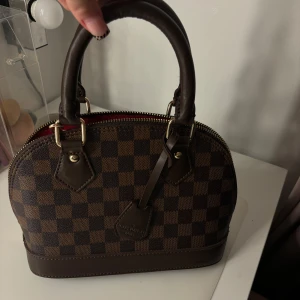 Brun rutig handväska - Säljer en snygg brun rutig handväska med dragkedja och röda detaljer inuti. Väskan har två robusta handtag och är perfekt för både vardag och fest. Den är i mycket bra skick och har en klassisk design som passar till det mesta. Kommer med band