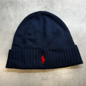  Polo Ralph Lauren mössa - Säljer en snygg mörkblå mössa från Polo Ralph Lauren. Den är stickad och perfekt för att hålla sig varm under höst och vinter. Mössan är i mycket bra skick och gjord av tvättbar merinoull. Passar perfekt till både vardag och fest! Den är ej använd och säljer den då jag ej använder den. 