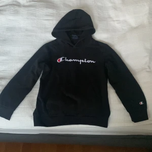 Svart hoodie från Champion - Säljer en svart hoodie från Champion i bra skick. Den har en klassisk Champion-logga på bröstet och en mindre logga på ärmen. Perfekt för höst och vinter, och den är supermjuk och bekväm. Passar både till vardags och chillkvällar. 