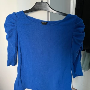 Blå topp från Lindex - Snygg blå topp från Lindex i storlek M. Den har trekvartsärmar med rynkade detaljer. 
