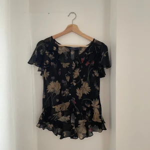 Blommig blus från Ralph Lauren - Säljer en elegant blommig blus från Ralph Lauren i svart med stora blommor i beige och inslag av rött och lila. Blusen har korta ärmar och volangdetaljer vid ärmslut och nederkant. Perfekt för både vardag och fest! 