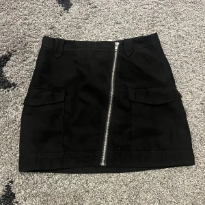 Svart kjol med dragkedja från Divided - Säljer en snygg svart kjol från Divided i storlek S. Kjolen har en cool dragkedja framtill och praktiska fickor på sidorna. Perfekt för både vardag och fest! 🖤